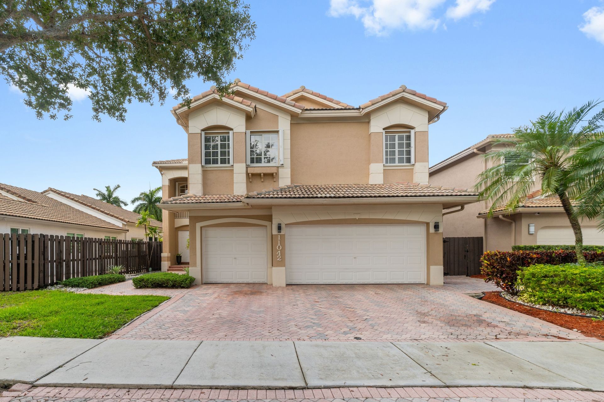 Two-story beige house with white garage doors, surrounded by trees and shrubs.